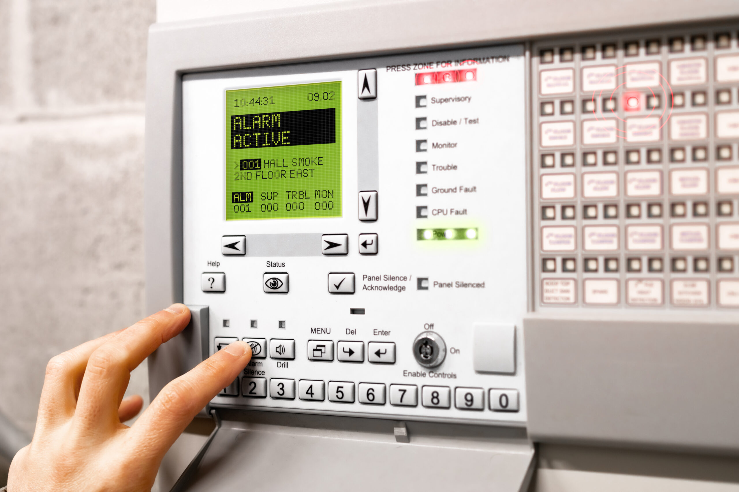 Fire alarm control panel is activated and in alert mode. Display message: Alarm active hall smoke. Red flickering lights and peeping. A hand is using the silence button. Selective focus.
